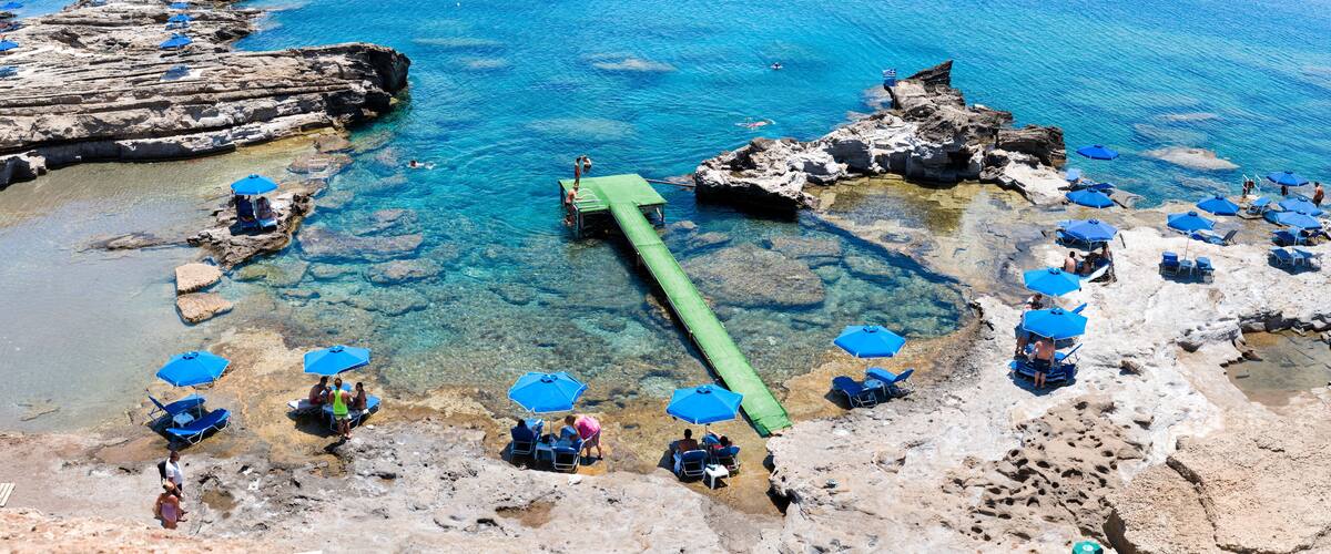A rocky beach with clear blue water, a green floating walkway, and blue umbrellas.