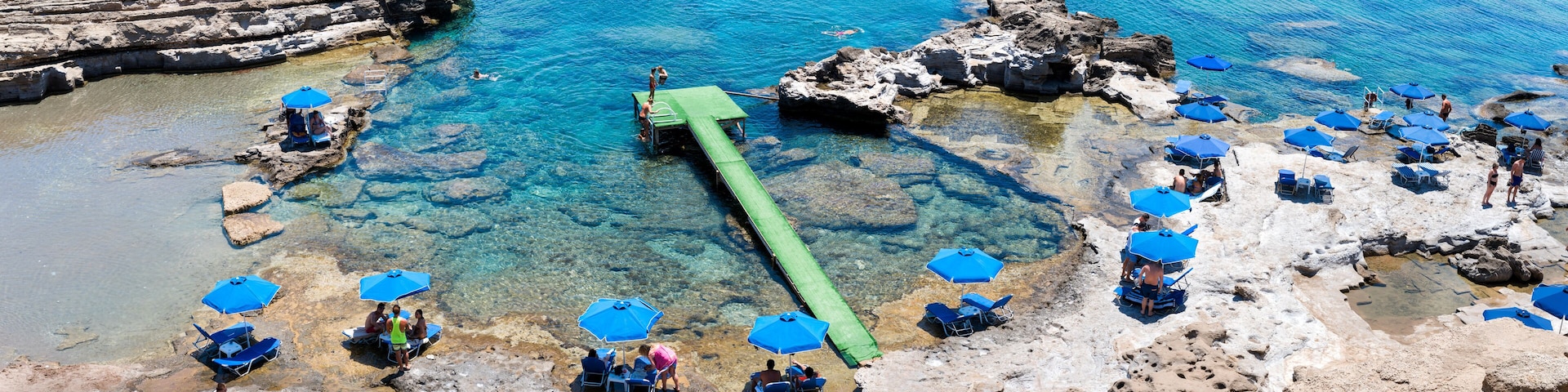 A rocky beach with clear blue water, a green floating walkway, and blue umbrellas.
