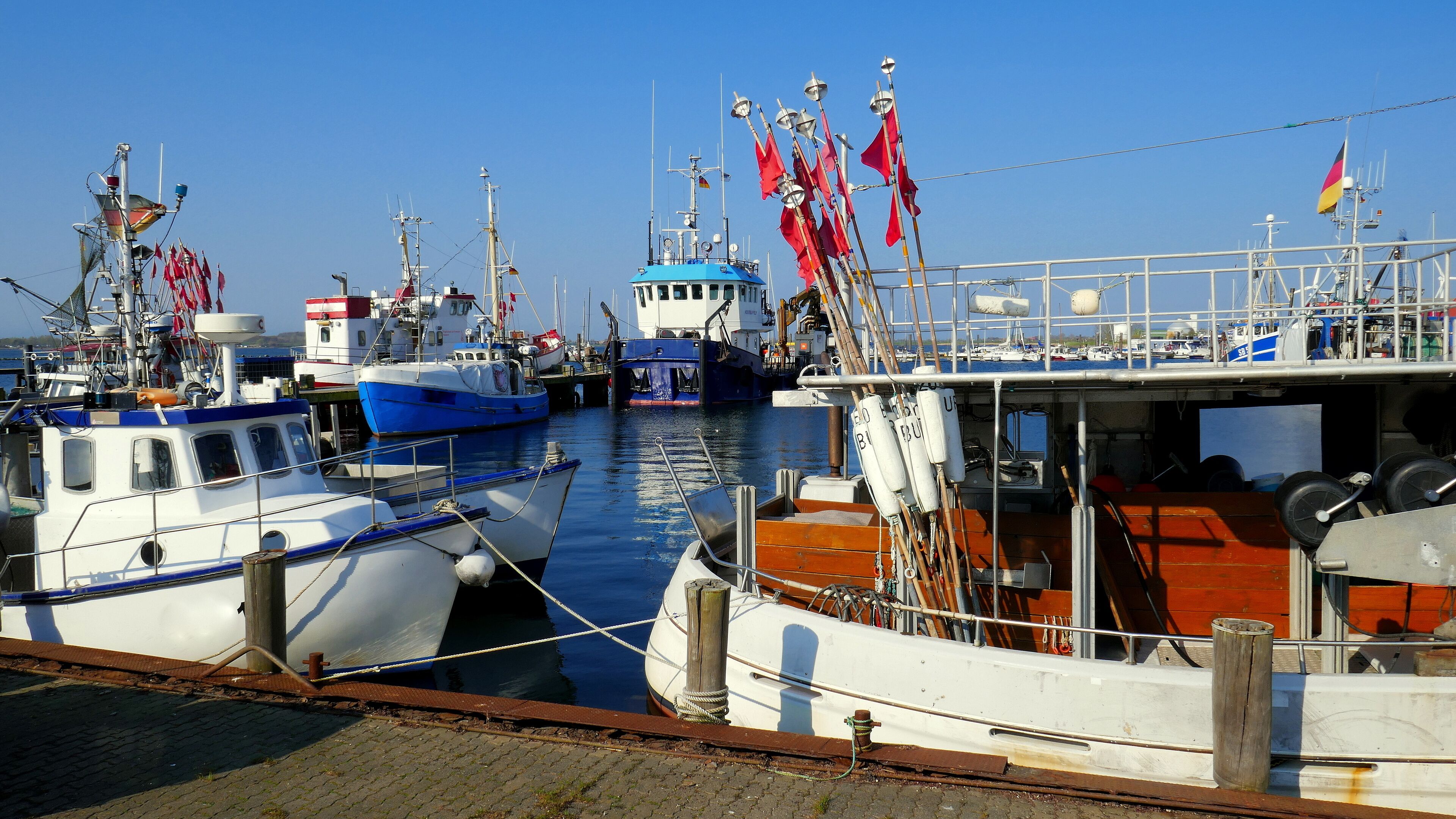 A marina with several boats docked, including a blue and white vessel and a white boat with red flags.