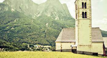 Uma igreja com cúpula vermelha e torre do relógio, cercada por um campo gramado e montanhas ao fundo.