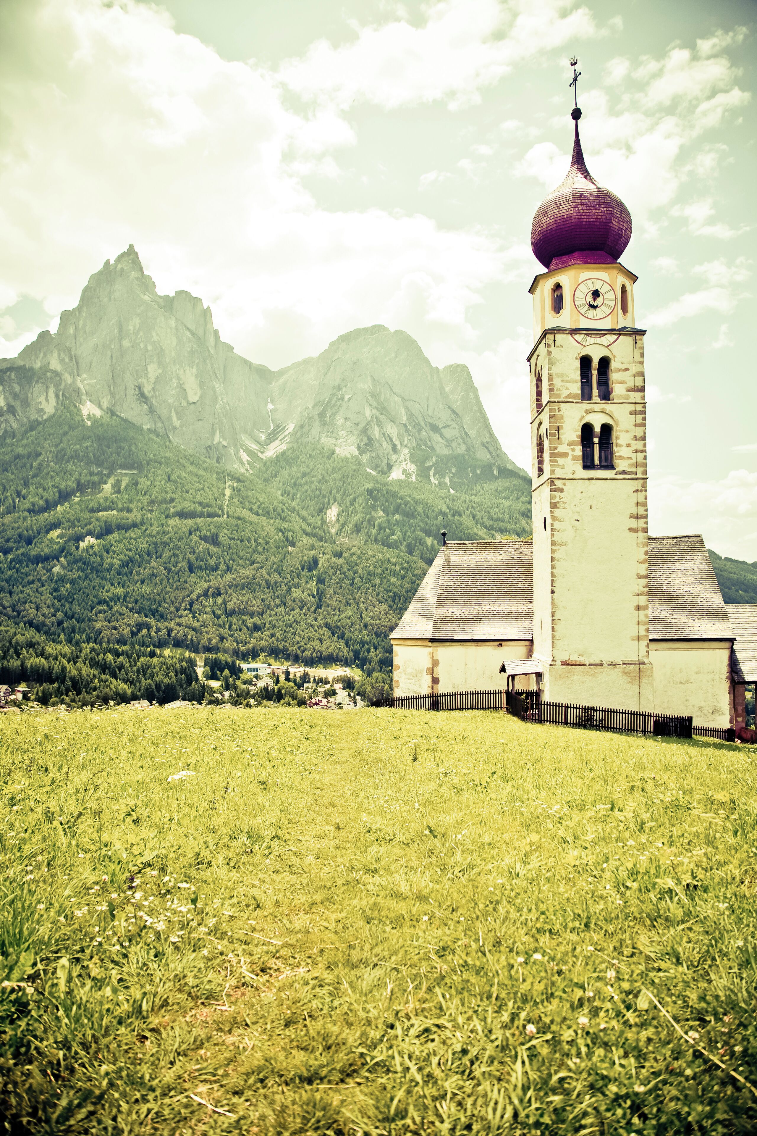 A church with a red dome and a clock tower, surrounded by a grassy field and mountains in the background.