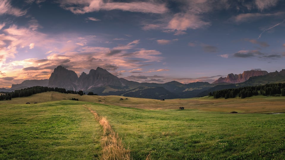 A grassy field with a small building, mountains, and a sunset sky.