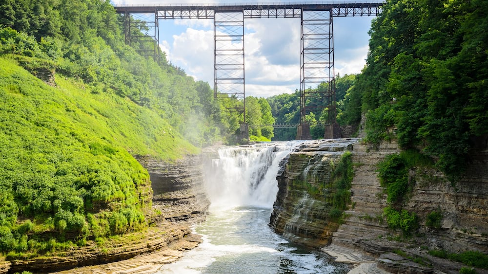 A waterfall cascading over a rocky cliff, a metal bridge spanning the river, and lush green hills.
