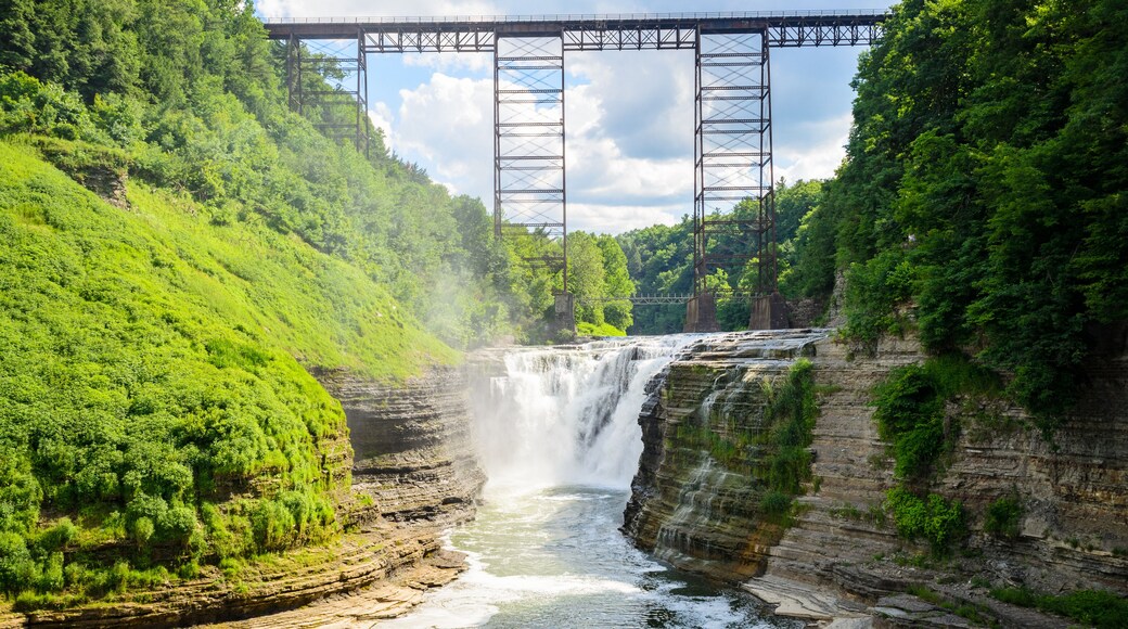 A waterfall cascading over a rocky cliff, a metal bridge spanning the river, and lush green hills.