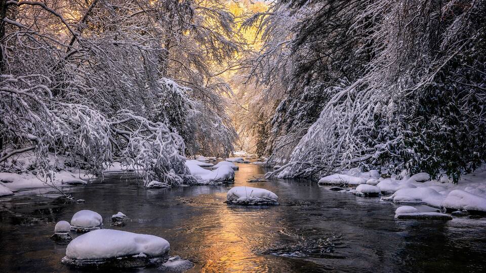 A snow-covered river with ice formations and trees reflecting sunlight.