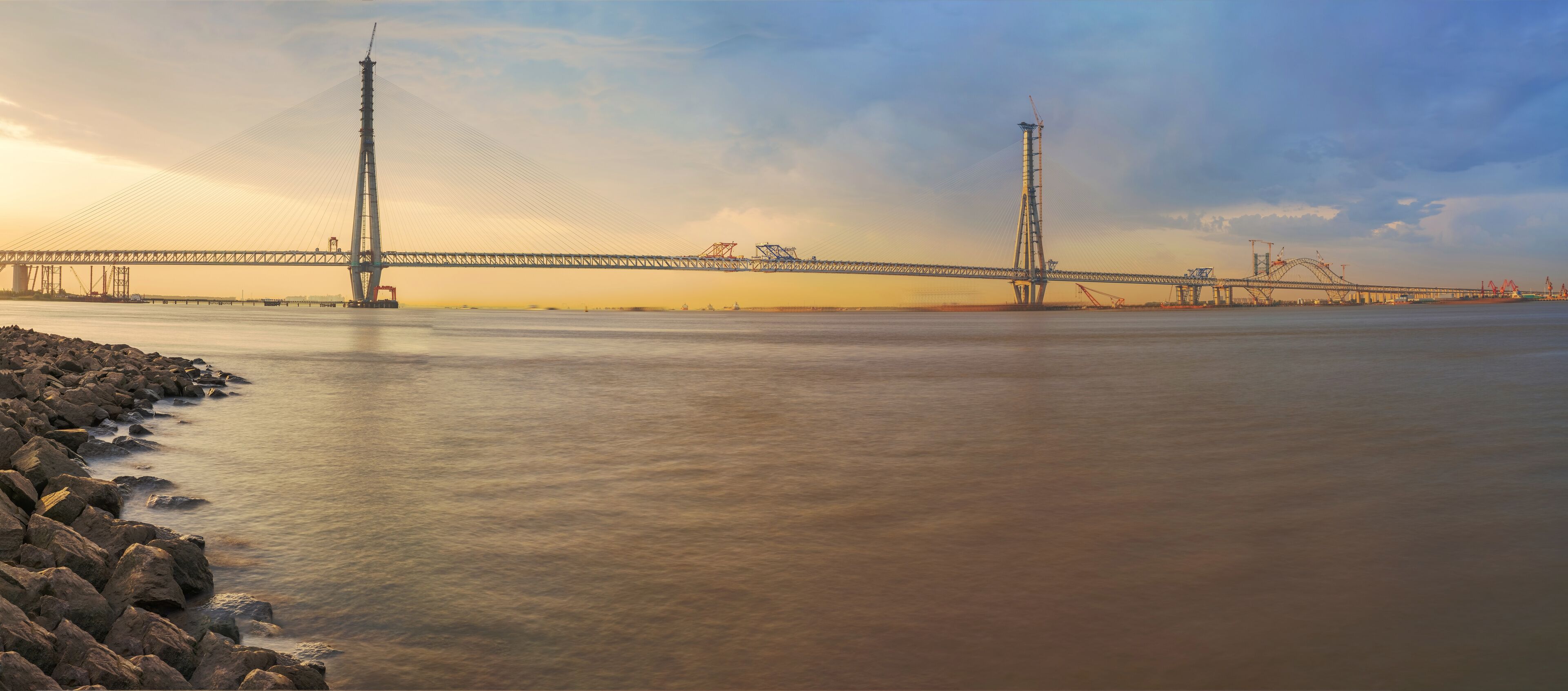 A large suspension bridge over a calm river with rocky shores and construction cranes in the background.
