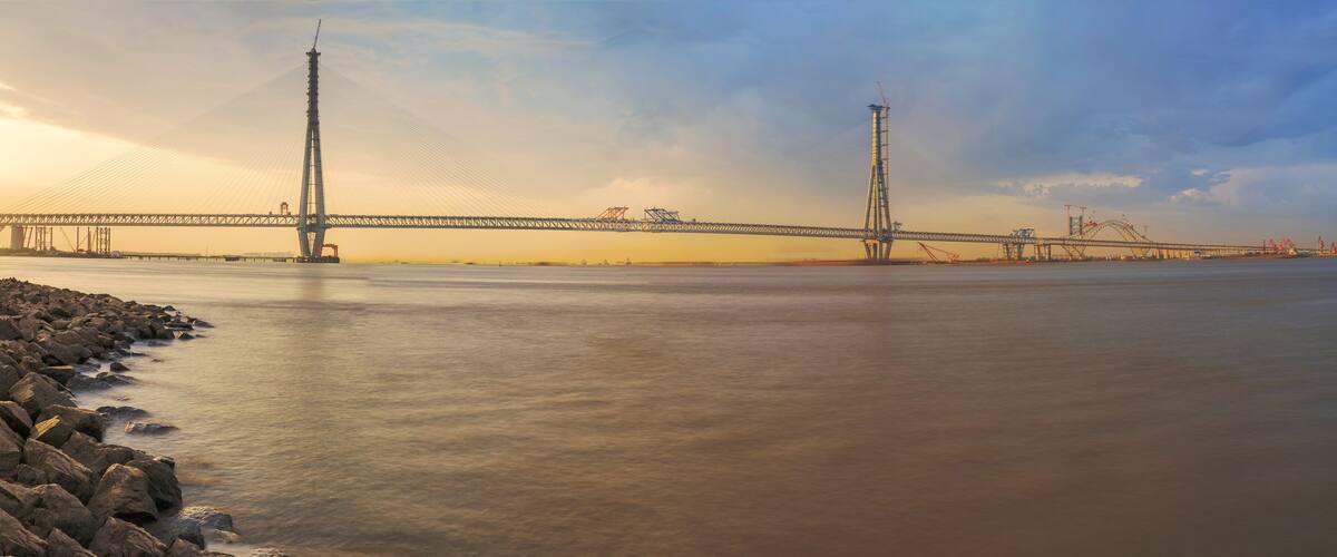A large suspension bridge over a calm river with rocky shores and construction cranes in the background.