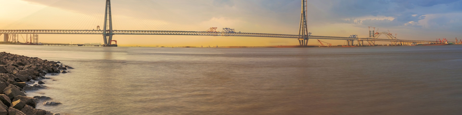 A large suspension bridge over a calm river with rocky shores and construction cranes in the background.