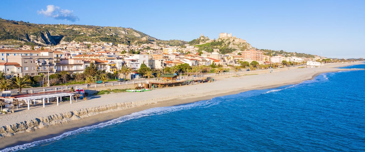 A coastal town with a beach, buildings, and a hill with a castle-like structure.