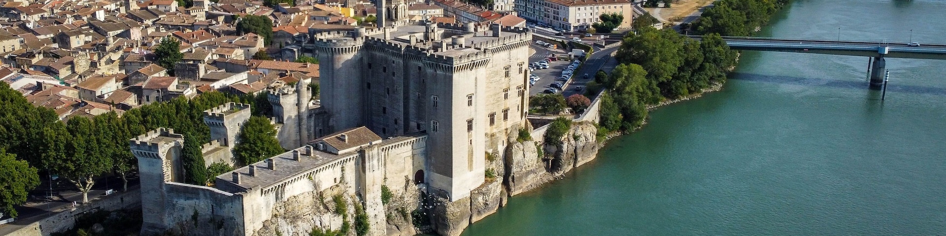 Un castillo medieval junto a un río, rodeado por un pueblo con un puente al fondo.