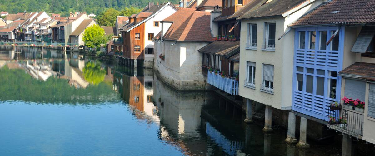 A calm river reflecting buildings, with a forested hill in the background.