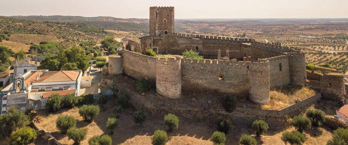 A medieval castle with a surrounding wall and a tower, set in a rural landscape with olive trees and distant hills.