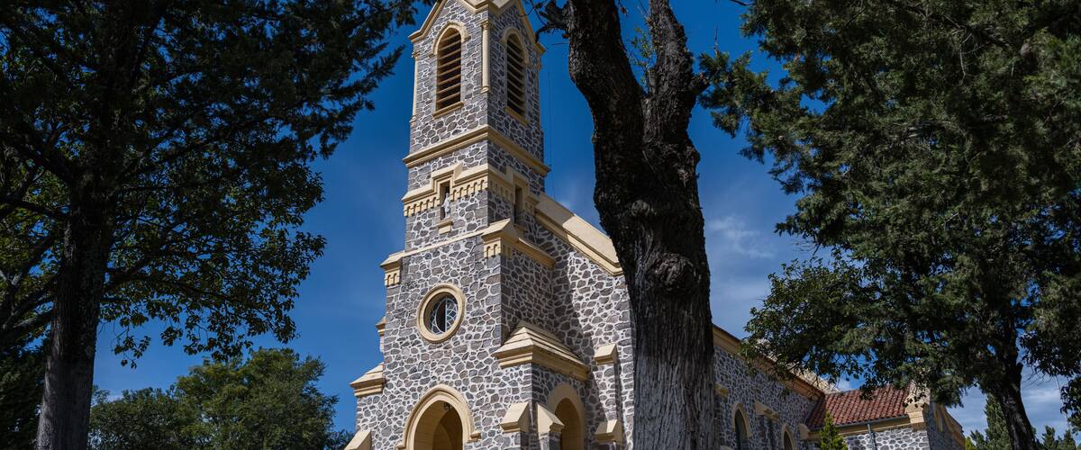A stone church with a bell tower, surrounded by large trees.