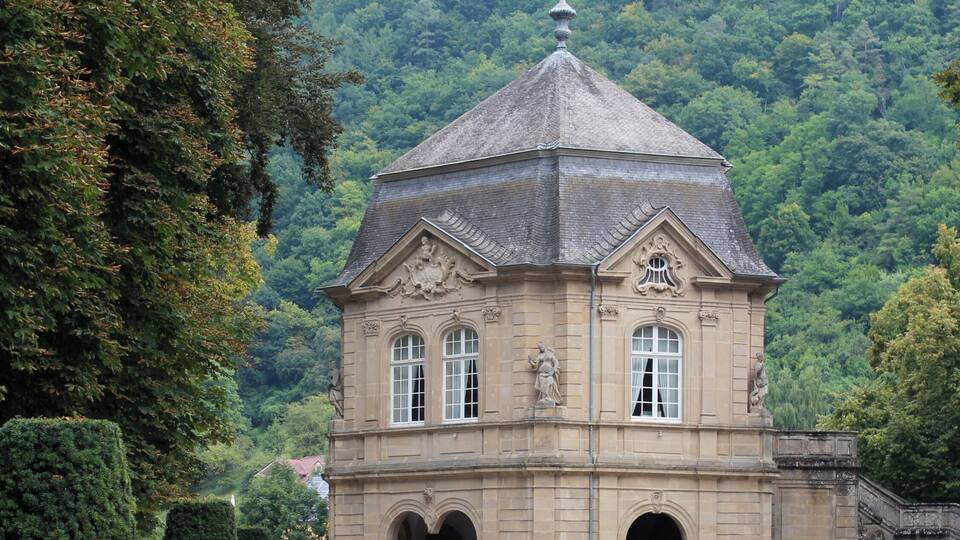 A historic building with a dome roof, surrounded by well-manicured gardens and a forested hillside.
