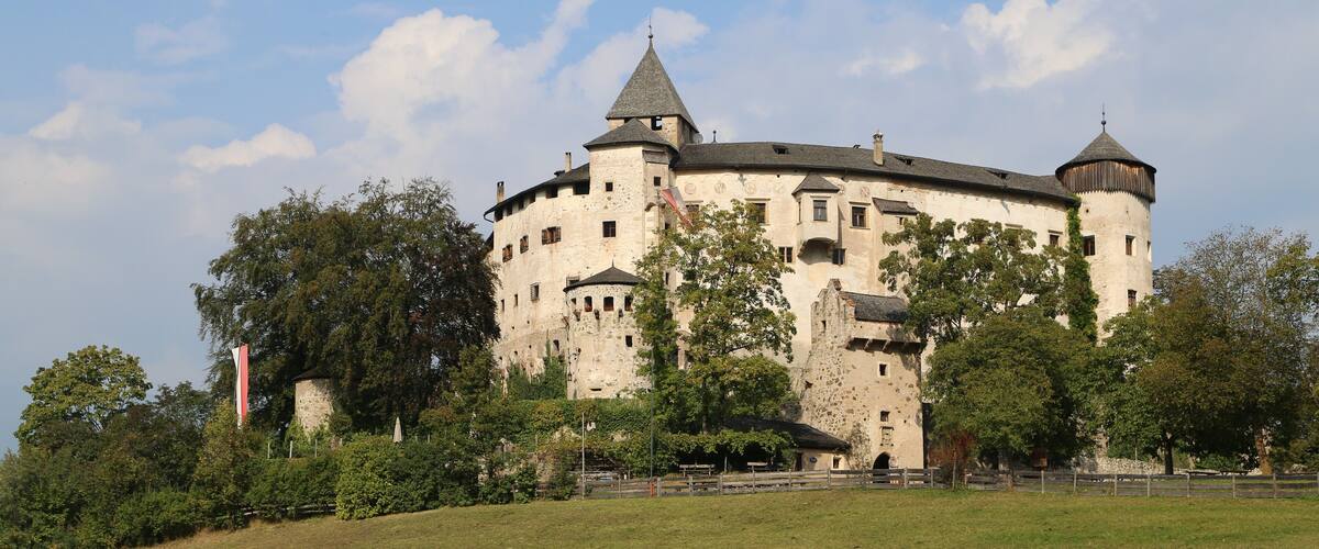 A historic castle with multiple towers and turrets, surrounded by lush greenery and a well-maintained lawn.