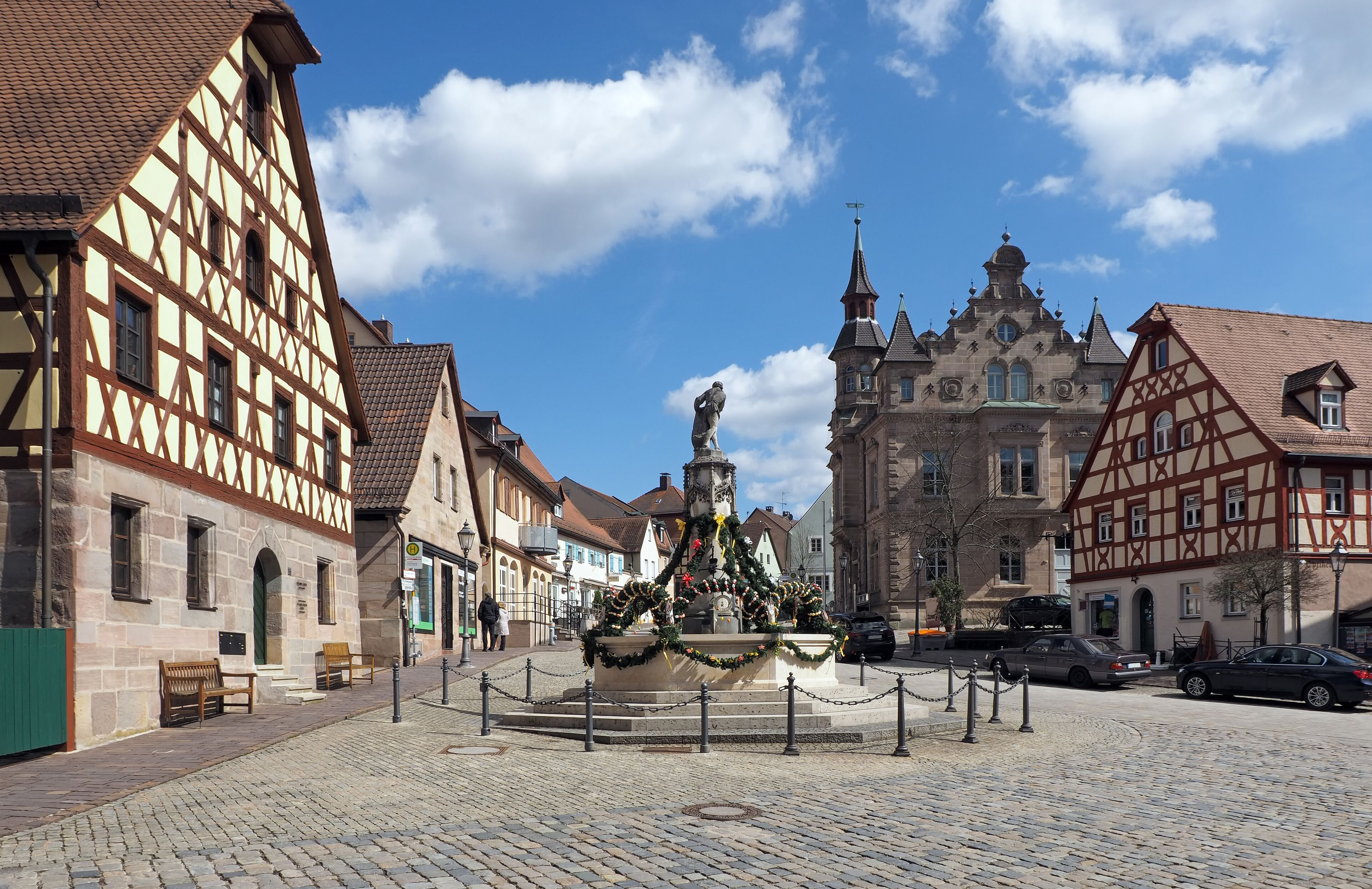 A cobblestone square with a fountain, traditional half-timbered buildings, and a church with a spire.