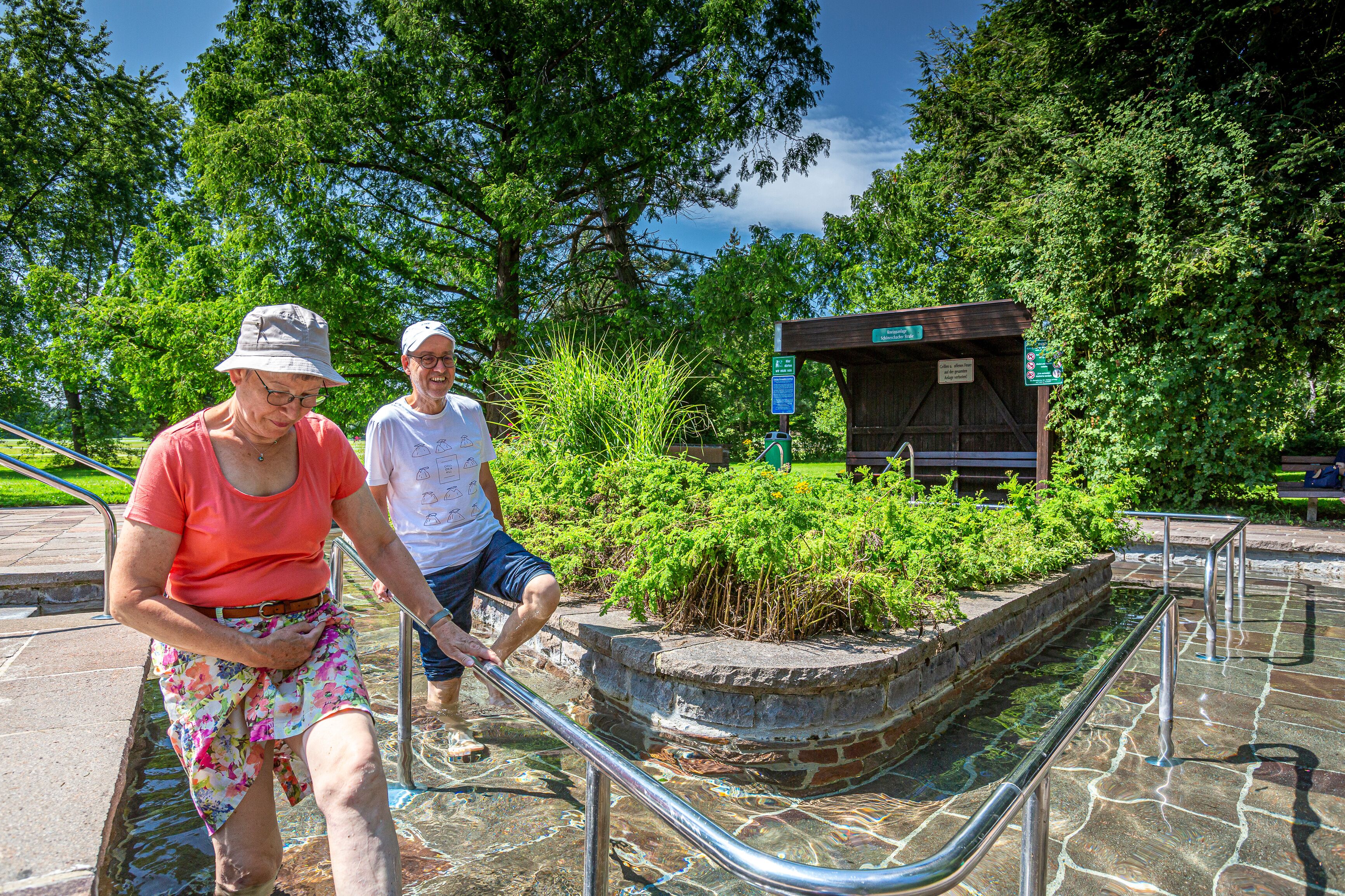 A man and a woman walking on a stone pathway with metal railings, surrounded by lush greenery and a wooden structure in the background.
