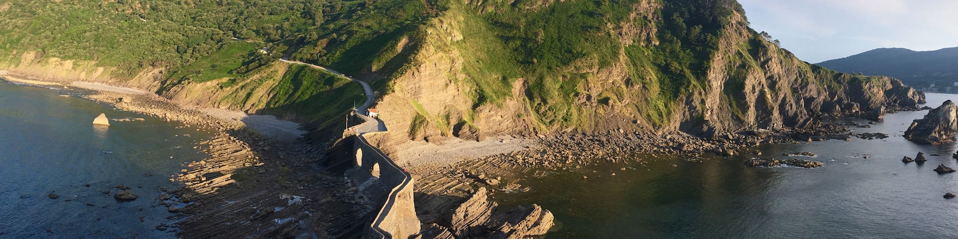 A coastal landscape with a winding road, rocky shoreline, and a hill with dense vegetation.