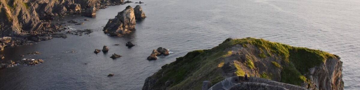 Eine Küstenlandschaft mit einem gewundenen Steinpfad entlang einer Klippe, der auf ein ruhiges Meer und ferne Hügel blickt.
