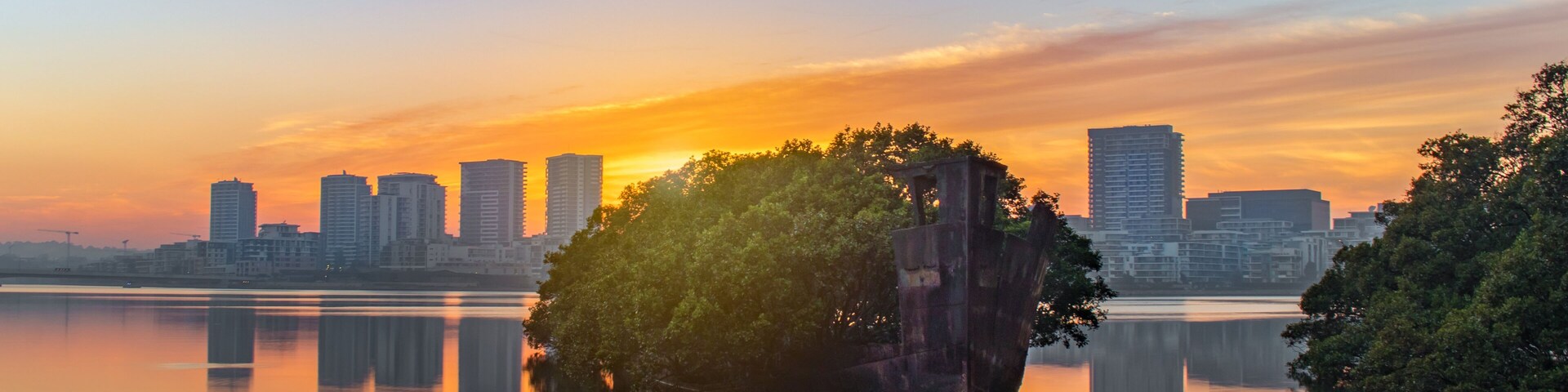 Horizonte urbano ao pôr do sol, com um corpo d'água tranquilo refletindo os edifícios e uma pequena ilha com árvores.