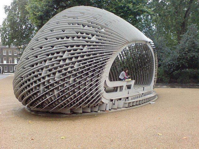 Uma estrutura esférica com design de treliça, uma pessoa sentada no interior e um edifício ao fundo.