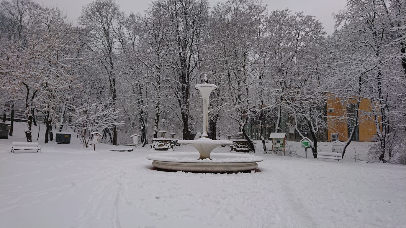 Un parque cubierto de nieve con una fuente central, árboles y un edificio amarillo al fondo.