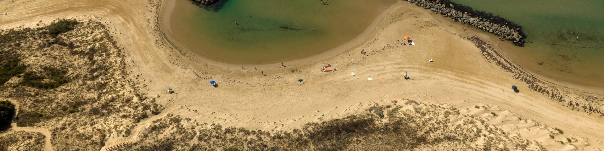 Aerial view of Donnalucata beach, a seaside resort in Sicily, Italy. The beach has fine sand, crystal-clear water, and is bordered by two stone breakwaters. It's a sunny summer day.