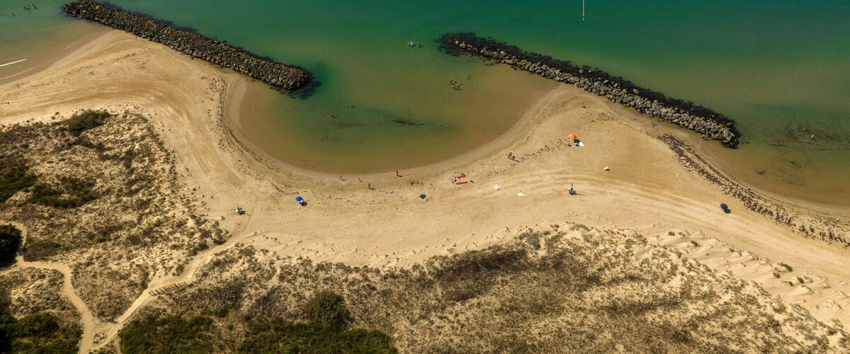 An aerial view of a beach with two breakwaters, clear turquoise water, and sparse vegetation.