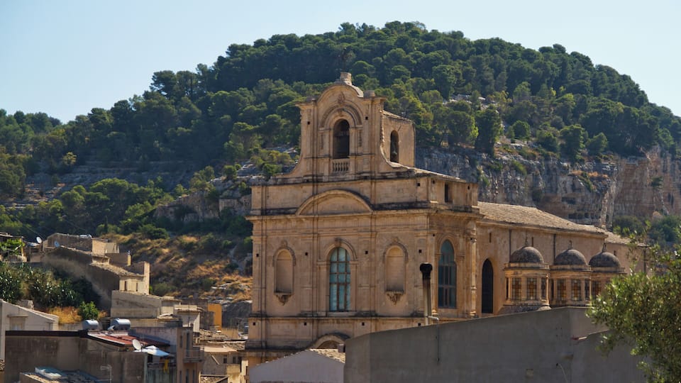 A historic church with a bell tower, set against a backdrop of a forested hill and a rocky cliff.