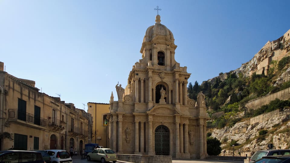 A historic church with a dome and statues, surrounded by old buildings and a rocky hillside.
