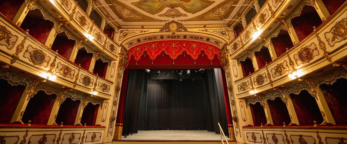 An ornate theater interior with a grand chandelier, red velvet curtains, and elaborate ceiling frescoes.