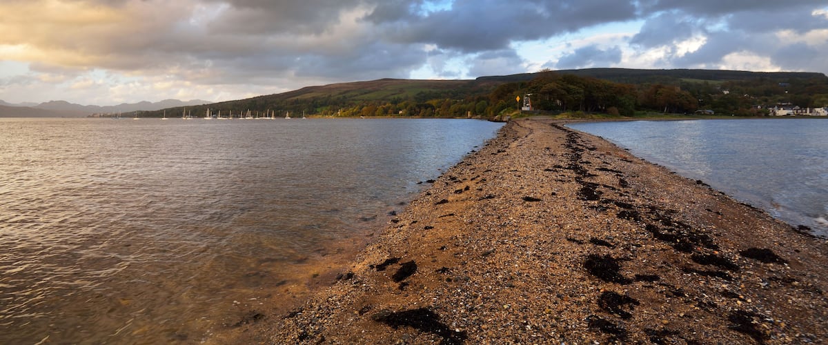 A pebbly beach with sailboats in the distance, a forested hill, and a cloudy sky.
