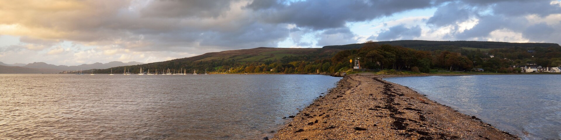 A pebbly beach with sailboats in the distance, a forested hill, and a cloudy sky.