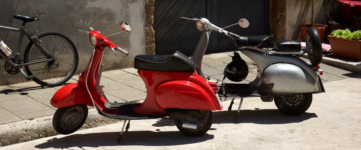 A red scooter parked on a sidewalk next to a building with a black door.