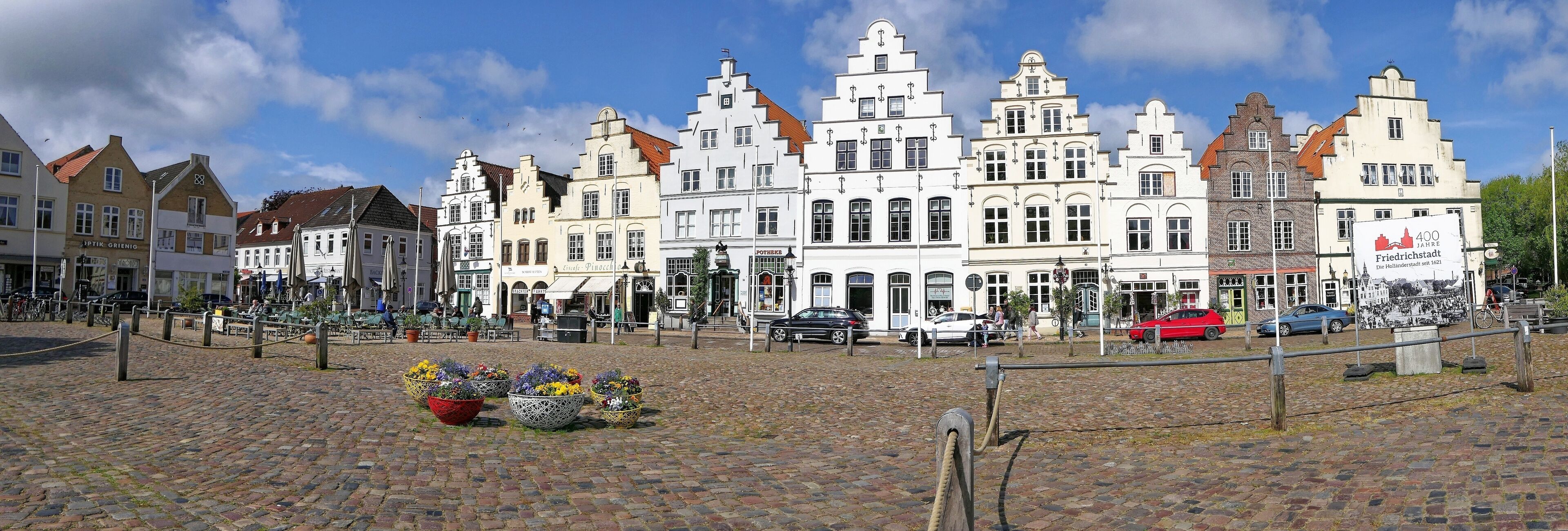 A cobblestone square with historic buildings, flower planters, and a sign indicating 400 years of Friedrichstadt.