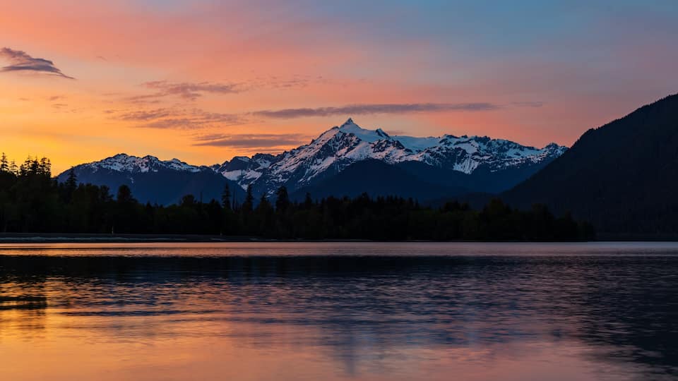 A mountain range with snow-capped peaks, a forested area, and a calm lake reflecting the sky.