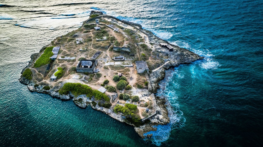 Vista aérea de una isla rocosa con estructuras históricas y aguas azul cristalino.