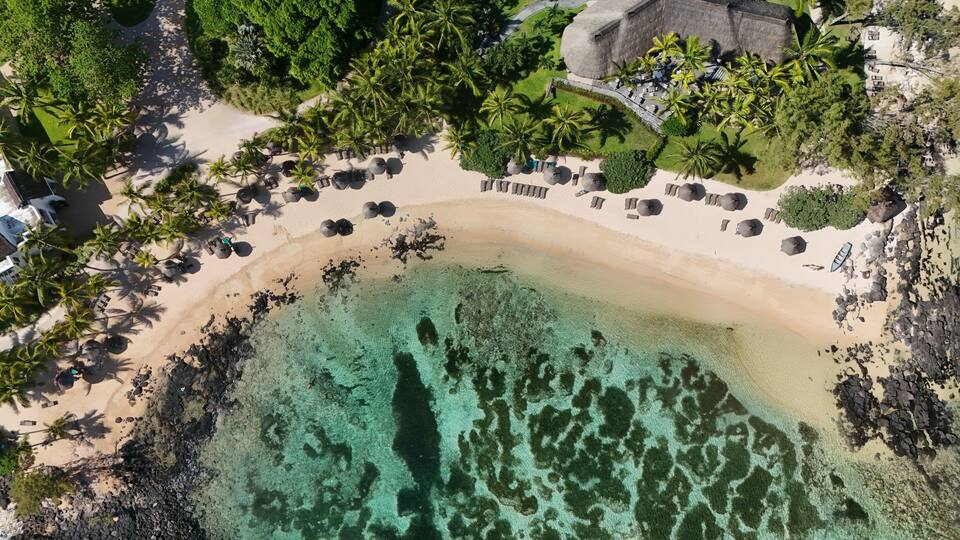 An aerial view of a tropical beach with clear turquoise waters, palm trees, and a thatched-roof building.