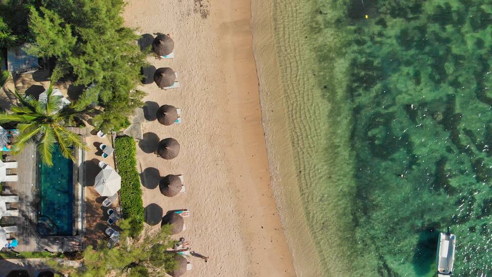 An aerial view of a beach with clear water, umbrellas, palm trees, and a pool.