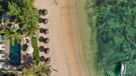 Eine Vogelperspektive eines Strandes mit klarem Wasser, Sonnenschirmen, Palmen und einem Pool.