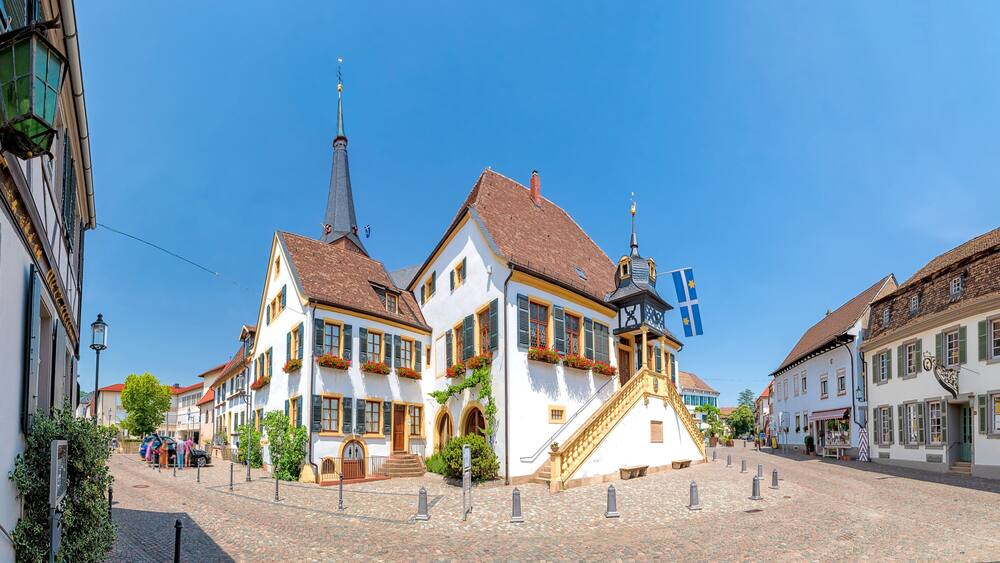 A cobblestone street lined with traditional European buildings, a church spire, and a flag.