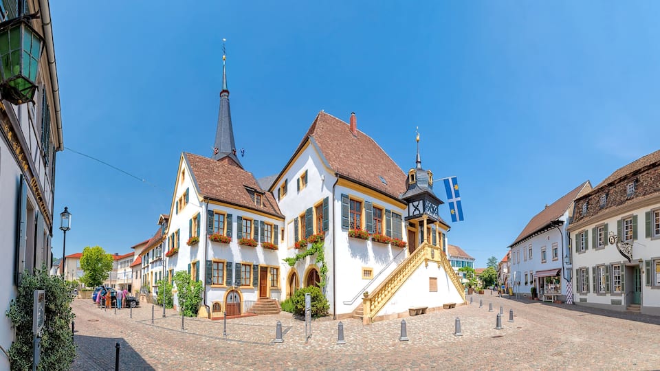 A cobblestone street lined with traditional European buildings, a church spire, and a flag.