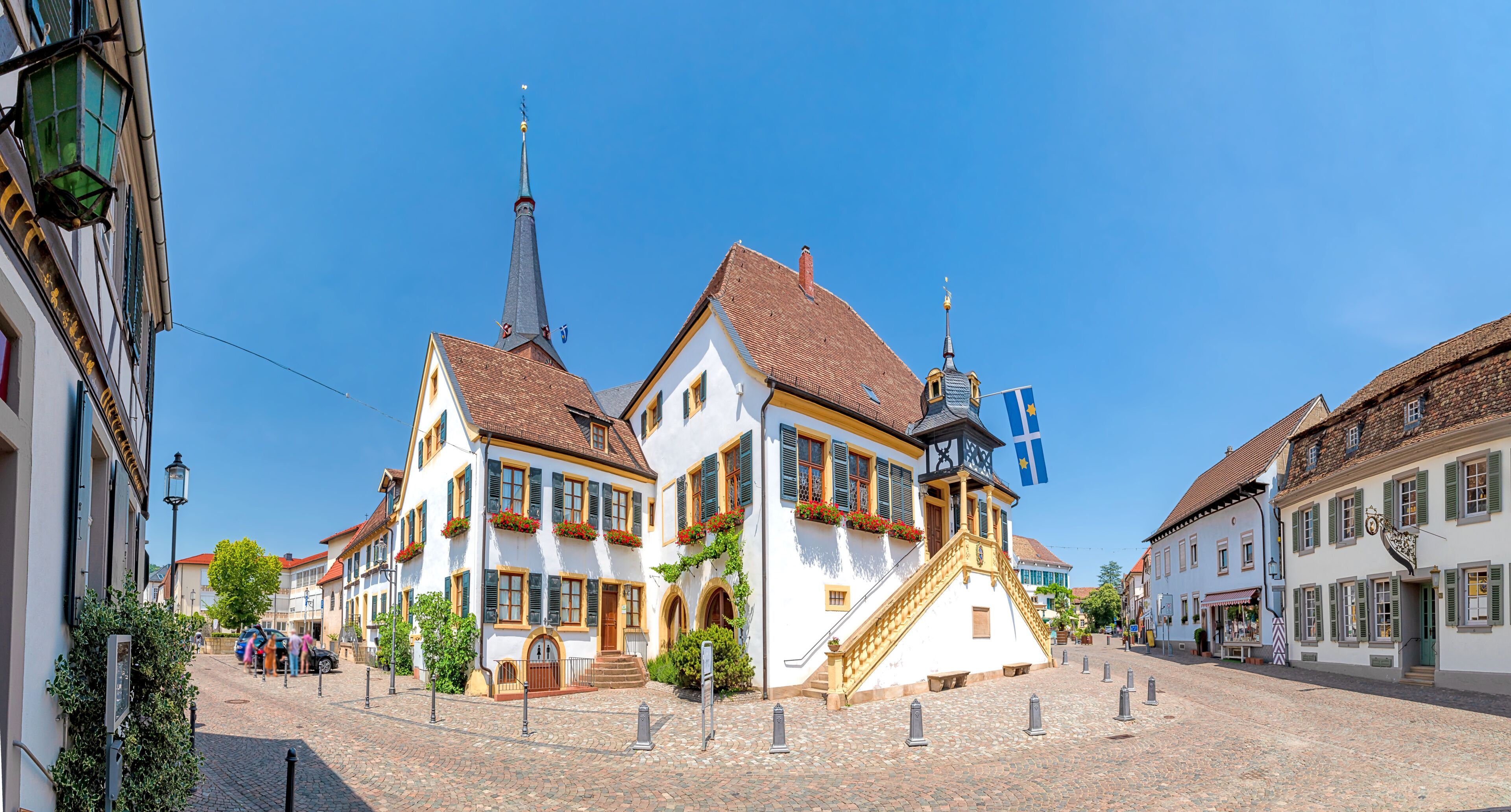 A cobblestone street lined with traditional European buildings, a church spire, and a flag.