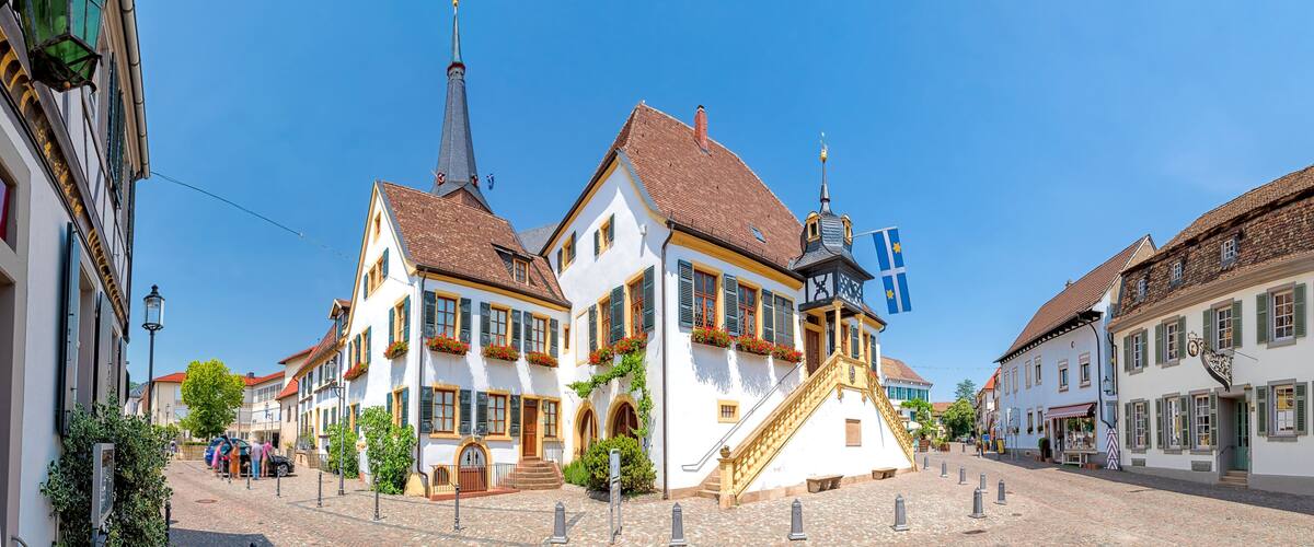 A cobblestone street lined with traditional European buildings, a church spire, and a flag.