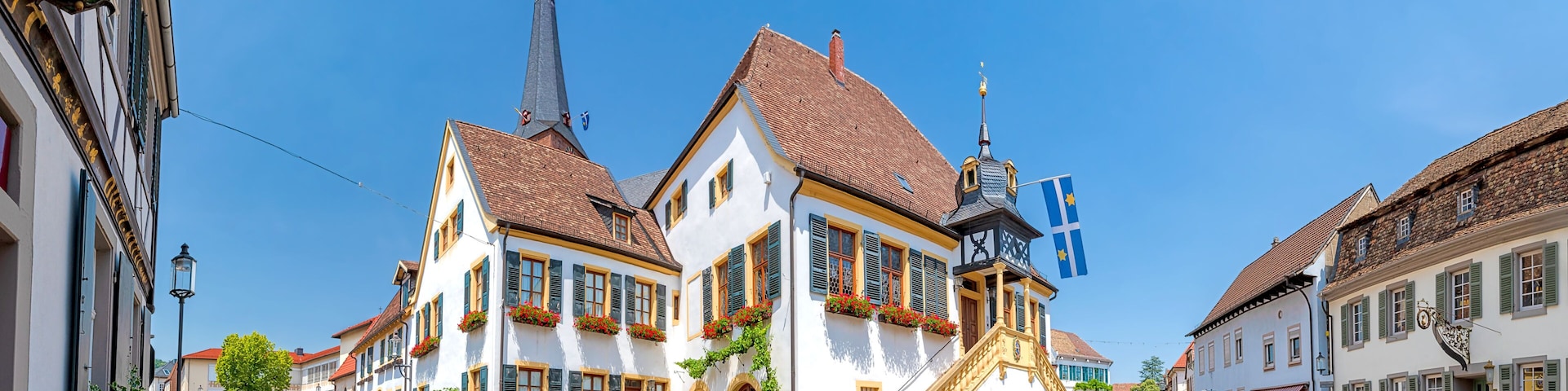 A cobblestone street lined with traditional European buildings, a church spire, and a flag.