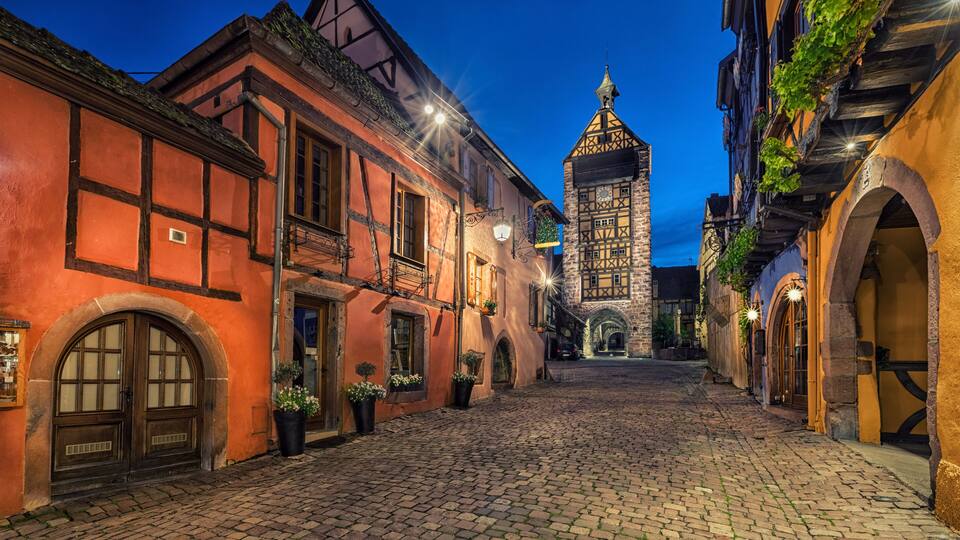 A cobblestone street lined with traditional half-timbered buildings and a prominent tower in the background.