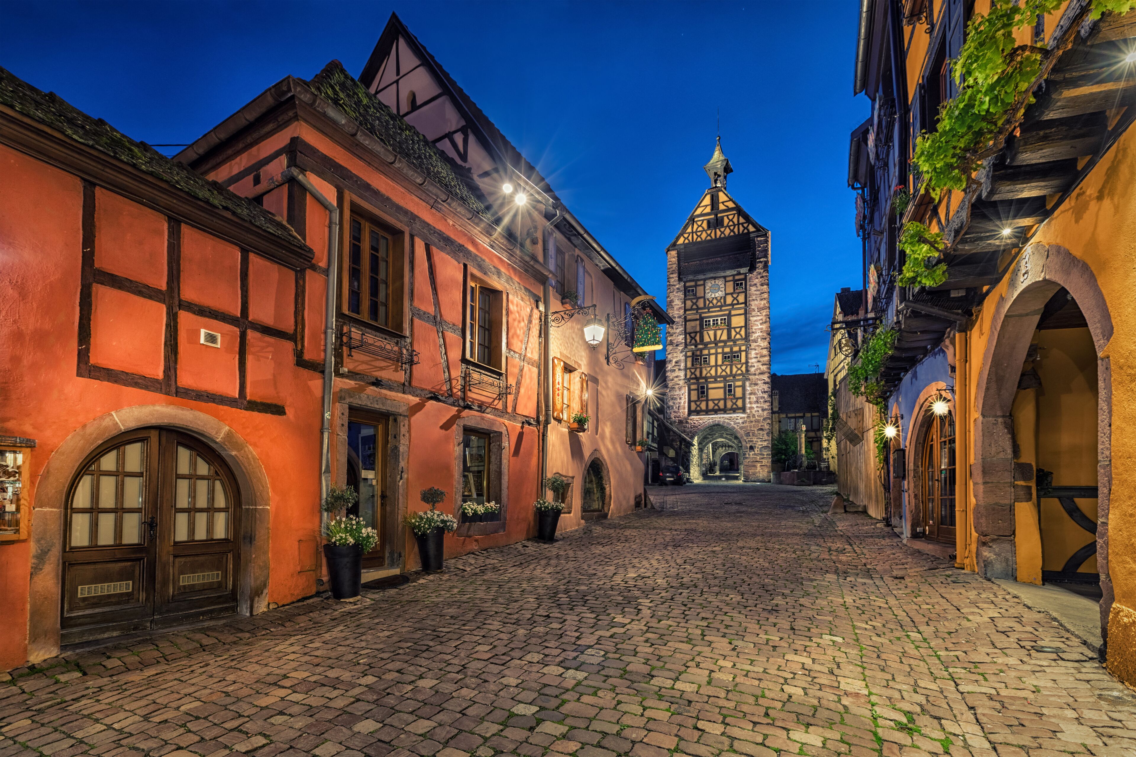 A cobblestone street lined with traditional half-timbered buildings and a prominent tower in the background.