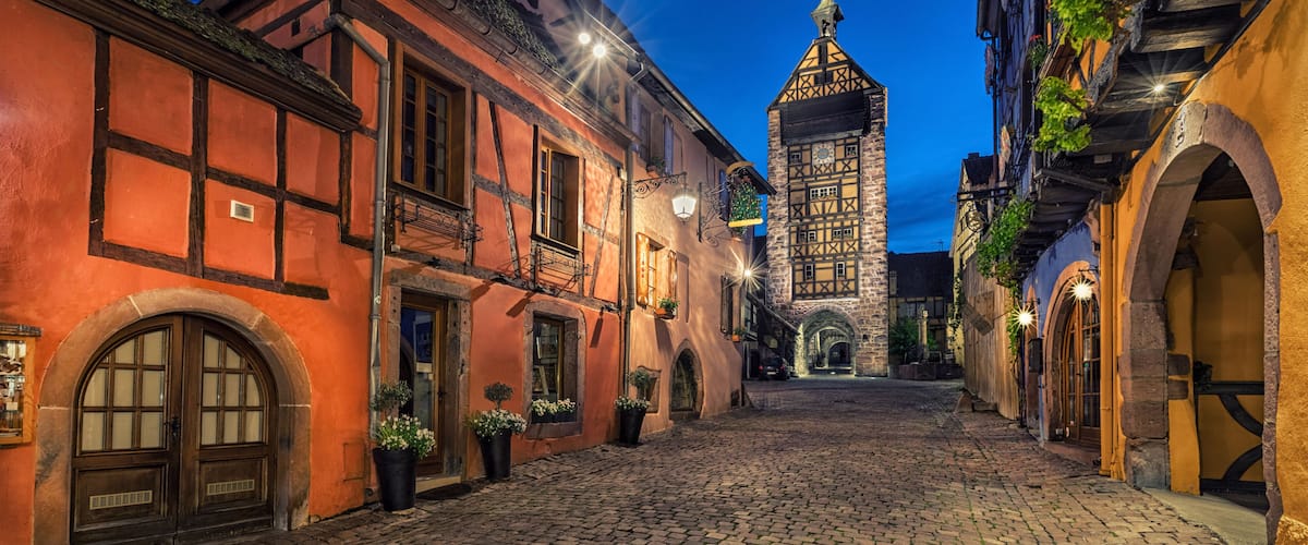 A cobblestone street lined with traditional half-timbered buildings and a prominent tower in the background.