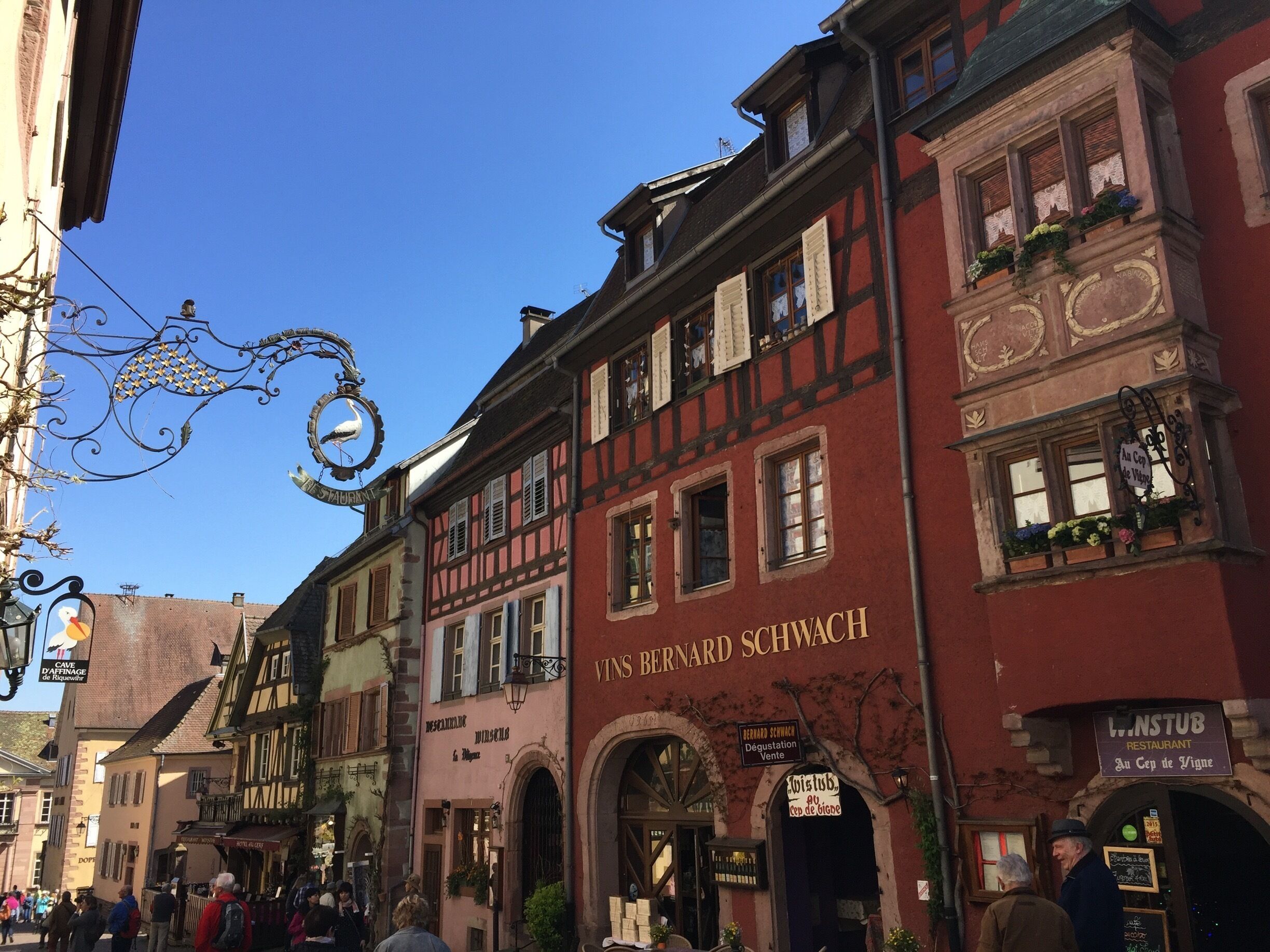 A street with traditional European architecture, including a building with 'VINS BERNARD SCHWACH' signage.