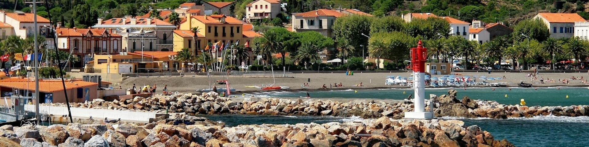 A coastal town with rocky breakwaters, a lighthouse, and terraced vineyards in the background.